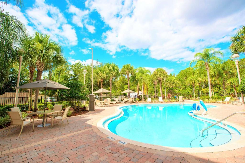 Outdoor swimming pool surrounded by palm trees, lounge chairs, and tables with umbrellas on a sunny day.