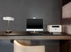 A modern desk setup with a computer monitor, keyboard, mouse, printer, and a desk lamp. The desk is against a dark textured wall, and a beige chair is in front of it.