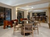 Modern dining area with round and rectangular tables, surrounded by cushioned chairs. Large windows on two sides. Soft lighting creates a welcoming atmosphere.