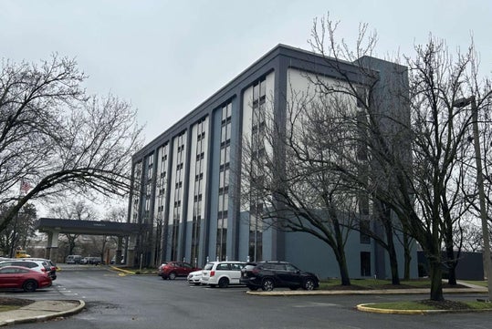 A six-story office building with a gray facade, surrounded by leafless trees and parked cars, under an overcast sky.