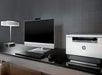A modern desk setup featuring a desktop computer with a widescreen monitor, keyboard, and mouse, alongside an HP printer and a sleek table lamp on a dark surface.