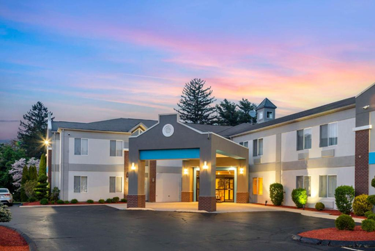 A two-story hotel exterior with illuminated entrance, surrounded by landscaped greenery and a sunset sky.