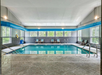 Indoor swimming pool with blue lane lines, surrounded by lounge chairs and windows. Bright overhead lighting and stone-tiled deck.