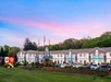 A two-story hotel with a Best Western Plus sign, surrounded by a parking lot filled with cars, green landscaping, and trees under a pink and blue sky.