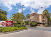 Exterior view of a Best Western Plus hotel with palm trees, landscaping, signage, and an American flag visible under a partly cloudy sky.