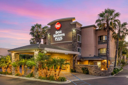 Exterior view of a Best Western Plus hotel at sunset, with palm trees, illuminated lights, and a covered entrance.