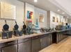 A hotel breakfast buffet counter features waffle makers, cereal dispensers, hot food trays, coffee machines, and decorative wall art above dark cabinets.