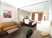 Hotel room with a double bed, sofa, desk, and TV. Walls have neutral tones, and a framed artwork is above the sofa. Natural light comes through a window with curtains.