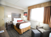 Hotel room with a neatly made bed, a nightstand with a lamp, an armchair, a small ottoman, and a large window with curtains.