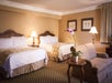 Hotel room with two double beds, floral artwork, a patterned armchair, and a small table with a potted orchid. Warm lighting from two bedside lamps.