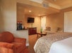 Cozy hotel room with a king-sized bed, a seating area featuring a sofa and armchair, a flat-screen TV on a dresser, and a coffee station. Beige and brown tones dominate the decor.