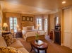 A spacious hotel room with a king-sized bed, sofa, coffee table with flowers, mini fridge, and cozy lighting. Decor features warm tones and framed artwork on the walls.