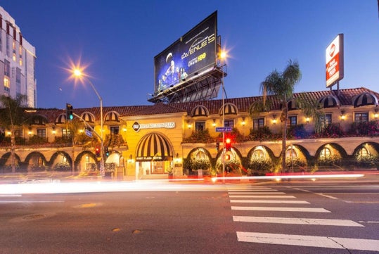 Street view of a hotel at night with a large billboard above, lit up windows, and streaks of car lights on the road. Crosswalk lines are visible in the foreground.