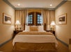 A cozy hotel room with a king-sized bed, flanked by two lamps on nightstands, curtained windows above, and framed art on beige walls.