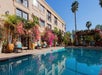 A poolside view of a three-story building with flowering plants, lounge chairs, and palm trees under a clear blue sky.