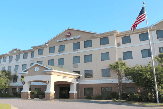 The exterior of the Best Western Plus Valdosta Hotel & Suites with a covered front entrance and trees in the landscaping on a sunny day.