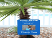 A blue sign for a dog park with a picture of a brown dog on it with a tree and some decorative rocks behind it.