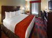 Hotel room with a large bed, white and red bedding, bedside lamps, an armchair, desk with a chair, a TV, and a window with curtains.