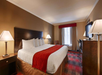 A hotel room with a large bed, red and white bedding, two bedside lamps, a television, a desk, and a window with curtains. The room has neutral walls and patterned carpet.