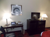 Hotel room interior with a desk, chair, and table lamp on the left. A framed spiral artwork is above. A TV on a dark cabinet with another lamp on the right. Carpeted floor, bins beneath the desk.
