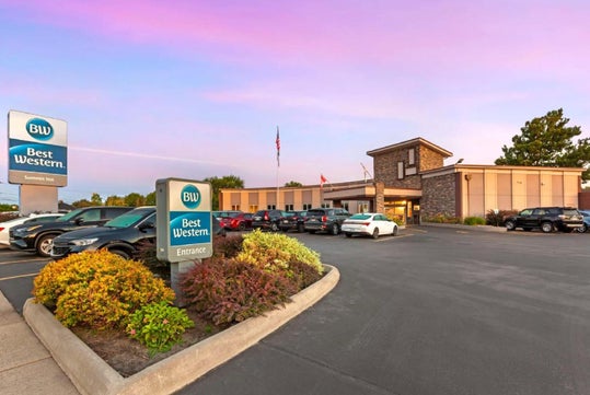 Exterior view of a Best Western hotel at sunset, with multiple parked cars, a driveway, signage, and an American flag.
