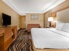 Hotel room with a large bed, brown sofa, wall-mounted TV on a wooden cabinet, and a patterned carpet. Walls are cream-colored with a framed artwork above the sofa.