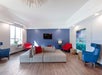 Modern lounge area with a gray sectional sofa, red and blue chairs, wall-mounted TV, and a beach-themed wall art. Light wood flooring, large windows, and overhead abstract light fixture.