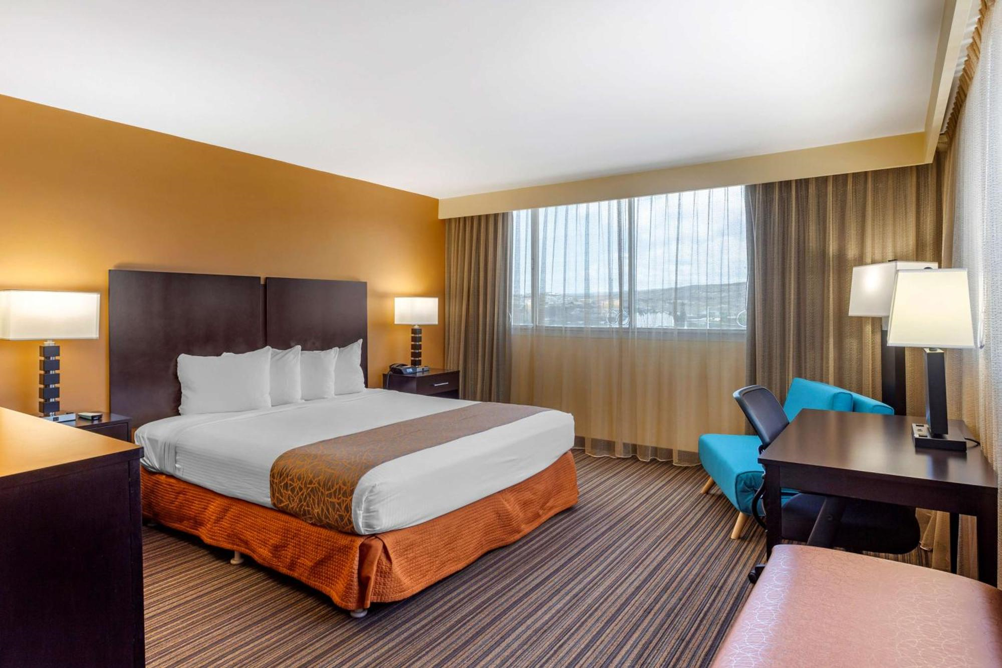 A hotel room with a double bed, two bedside tables with lamps, a desk with a chair and lamp, an armchair, and a large window with curtains.