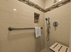 Accessible walk-in shower with beige tiles, a built-in shelf, a grab bar with a towel, a handheld showerhead, and a foldable wooden shower seat.