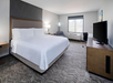 Hotel room with a king-sized bed, armchair, desk, TV, and window. Room features neutral tones and modern decor.