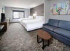 Hotel room with two double beds, a gray sofa, a wooden table, a TV, and a desk. Walls are light-colored with two abstract paintings. Carpet flooring and a large window with closed blinds.