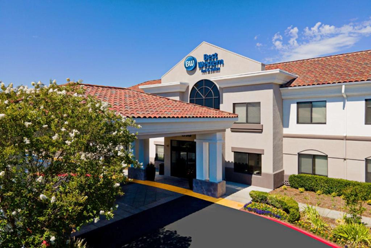 Exterior view of a Best Western Hotel with a red-tiled roof, white walls, and a sign above the entrance. Blue sky and greenery surround the building.