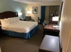 Hotel room with a large bed, lamps, armchair, desk, chair, and TV on a cabinet. Walls are light and teal, with a framed picture. Carpeted floor and curtains on the window.