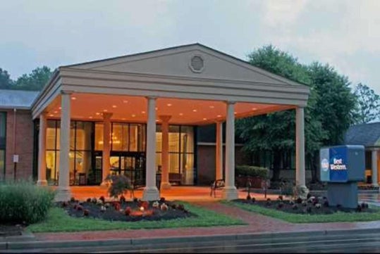 Front view of a hotel entrance with large columns, a lit interior, and nearby landscaping. The Best Western sign is visible on the right.