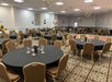A banquet hall with round tables covered in black tablecloths, set with glasses and decorations. Brown chairs surround the tables. The room is decorated with balloons.