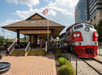Branson Scenic Railway station with an American flag flying. A vintage red and white train with the number 98 is parked on the track. People are gathered near the station entrance.