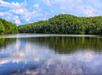 A calm lake reflects surrounding green trees and cloudy blue sky, with a wooded hill in the background.