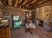 A rustic log cabin interior with a green wood stove, wooden table, and stone fireplace, featuring an exposed beam ceiling and wooden shelves.