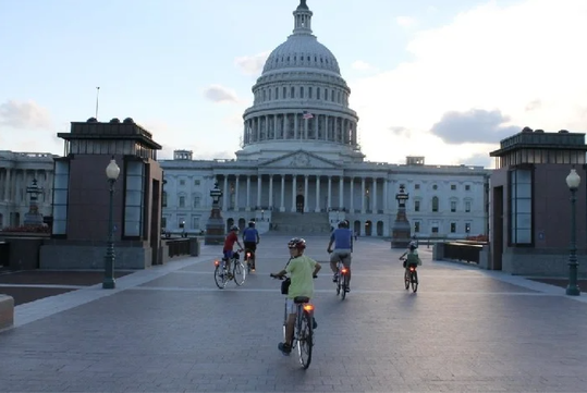 Cyclists riding towards the United States Capitol building under a partly cloudy sky.