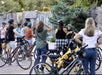 A group of people with bicycles, some wearing helmets, gathers outdoors near a stone wall and waterfall, listening to a person speaking.