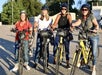 Four women wearing helmets stand outdoors with bicycles, smiling at the camera. The scene is sunny with trees and buildings in the background.