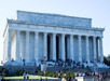 Lincoln Memorial with people gathered on steps, surrounded by greenery, under a clear blue sky.