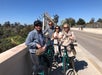 Four people with bicycles stand on a sunny bridge, smiling at the camera. Trees and a building are visible in the background.