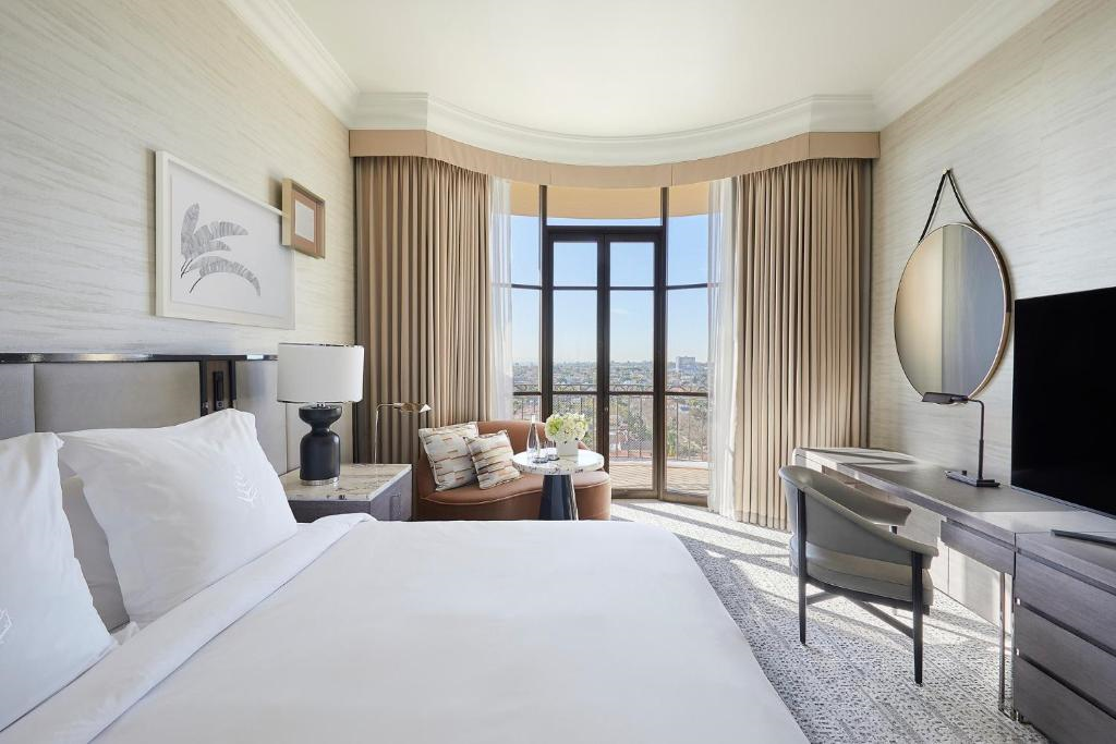 Modern hotel room with a bed, side table, armchairs by a large window, desk with a TV, and a round mirror. Neutral tones and city view through the window.