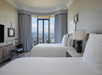 Hotel room with two double beds, a TV, desk, and large window with city and mountain view. Neutral decor with beige walls and curtains. Lamp on bedside table.