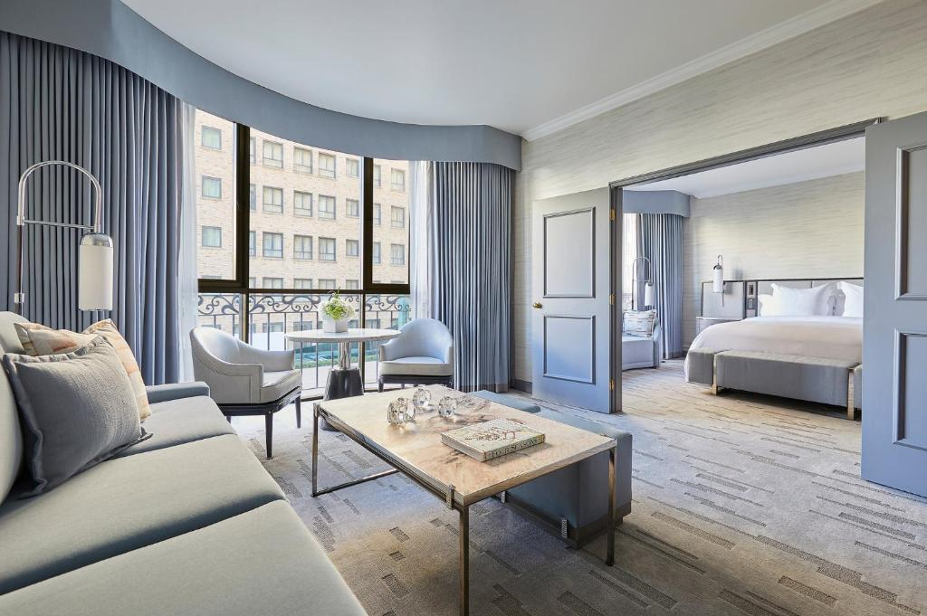 A modern hotel room features a seating area with a sofa, two chairs, and a coffee table. The bedroom with a bed is visible through an open door. Large windows and curtains complete the decor.