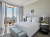 Hotel room with a king-sized bed, a small round table and chair, and an outdoor balcony. The room has neutral tones with a scenic city view through large windows.