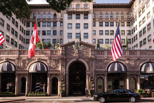 Exterior view of a grand hotel with ornate architecture. Canadian and American flags are displayed at the entrance. A luxury car is parked in front of the building under a clear sky.