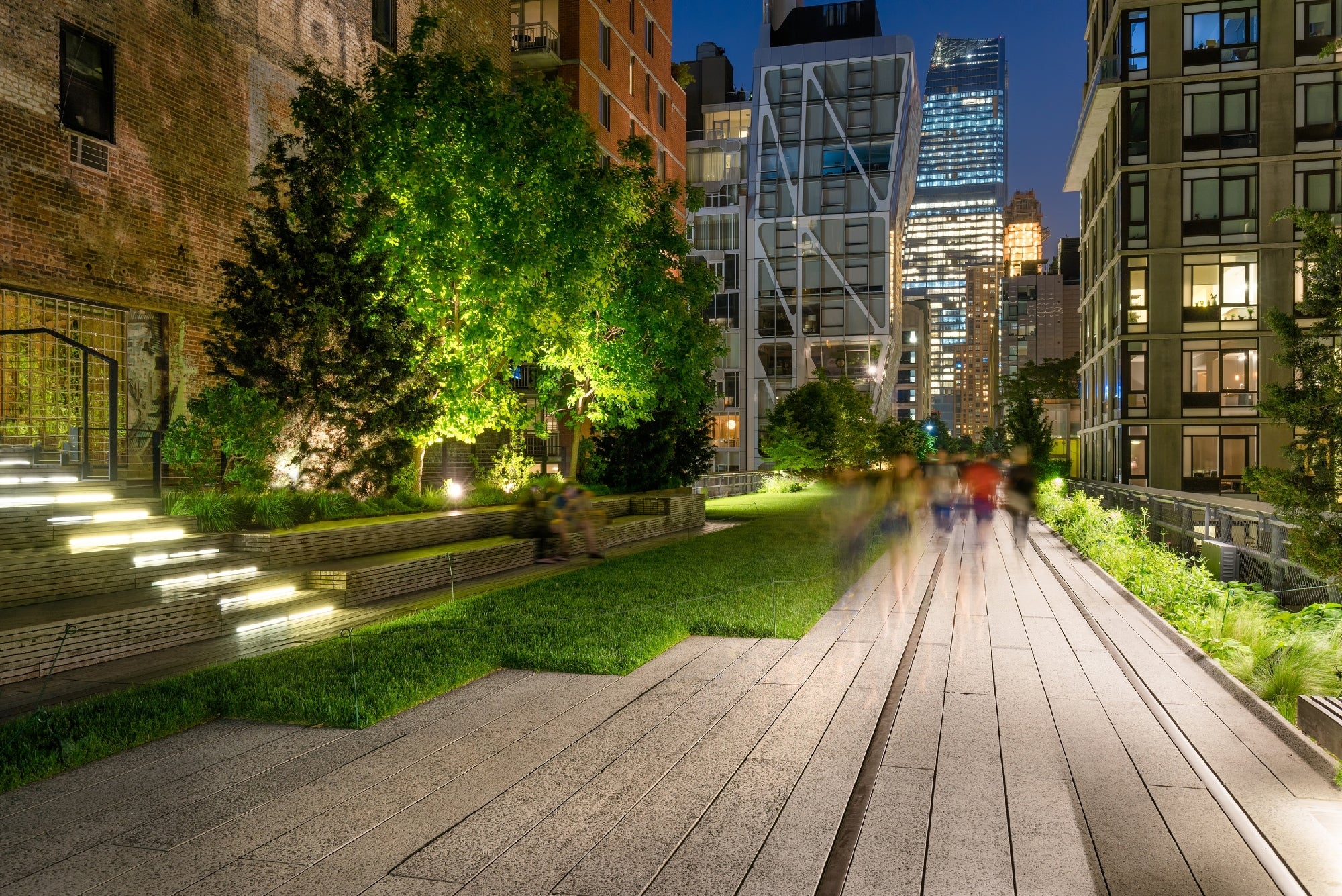 Urban park at night with modern architecture and illuminated pathway. Blurred figures walk along the path, surrounded by trees and buildings in the cityscape.