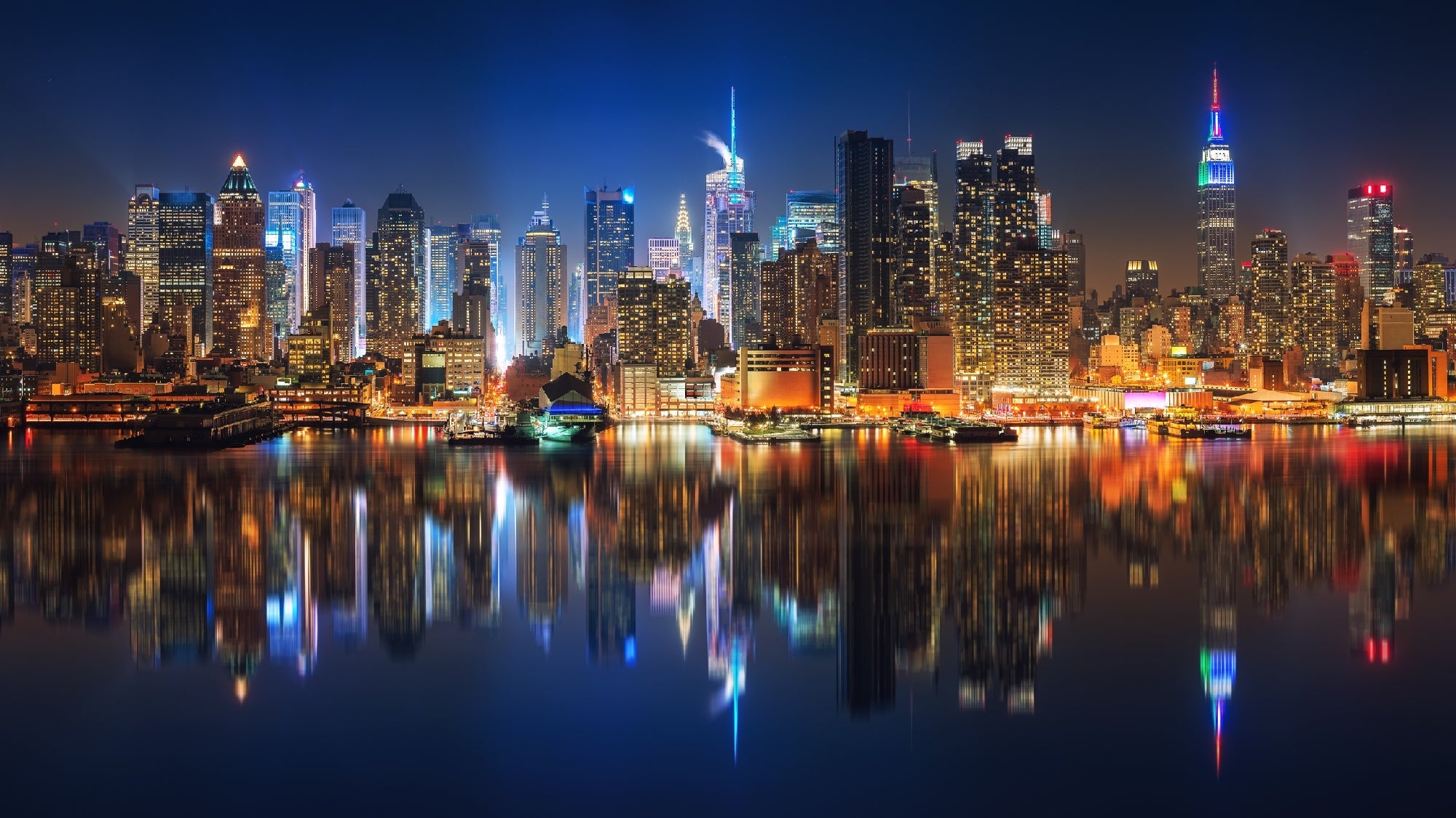 Nighttime cityscape of a brightly lit skyline with skyscrapers reflecting on a calm body of water.